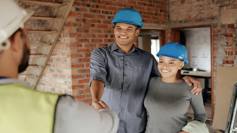 Un couple portant des casques de sécurité serre la main d’un professionnel sur un chantier, illustrant la confiance et la collaboration dans le choix d’une entreprise de rénovation.