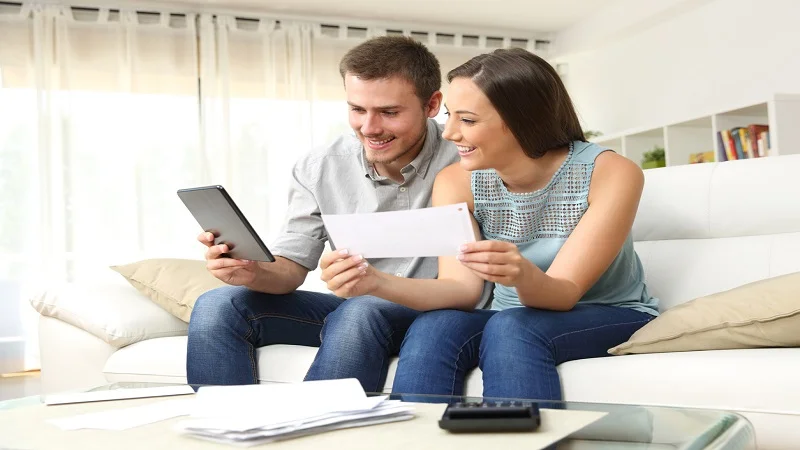 Un couple souriant est assis sur un canapé, examinant une lettre et consultant une tablette, ils sont en train de comparer des devis ou de gérer leur budget ensemble.