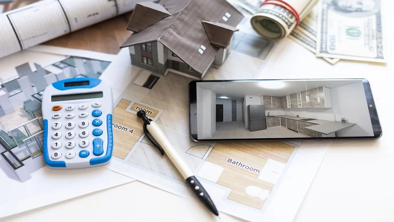 La planification financière d’un projet de rénovation de maison, avec des plans architecturaux, une calculatrice, un stylo, une maquette de maison, un smartphone affichant une modélisation 3D d’une cuisine rénovée, ainsi que des billets de banque et des rouleaux de dollars, symbolisant le budget et le financement des travaux.