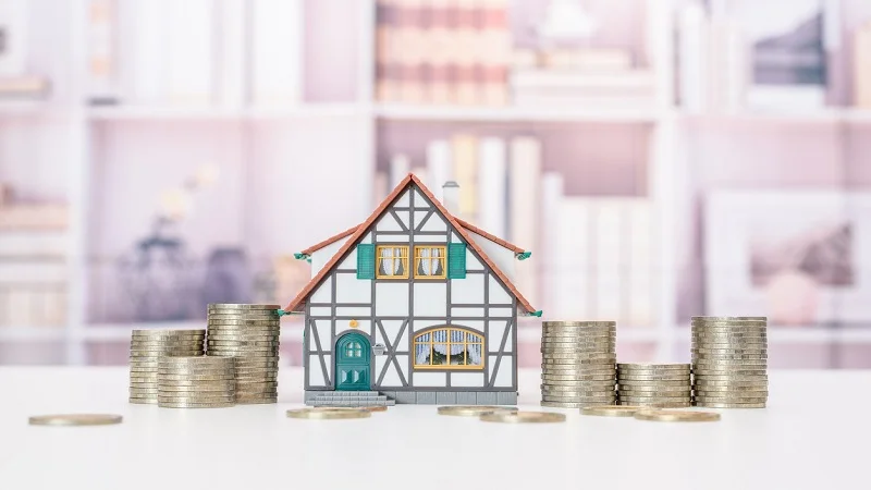 Une maquette de maison entourée de piles de pièces de monnaie, symbolisant les coûts ou l'investissement financier lié à la rénovation.