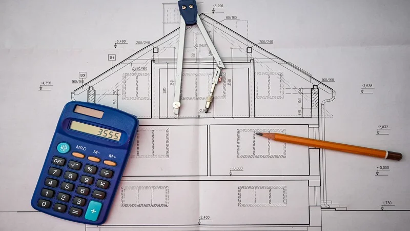 La planification d’une rénovation de maison, avec un plan architectural détaillé, une calculatrice, un compas et un crayon, symbolisant le calcul des coûts, la conception des travaux et l’élaboration du budget de rénovation.