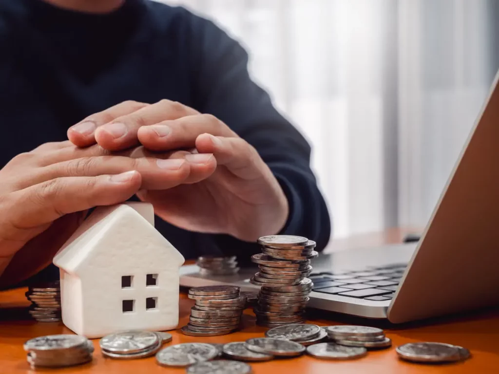 Vue sur une petite maison, à côté de plusieurs pièces de monnaie et abritée par deux mains pour faire référence à la rénovation maison aide.