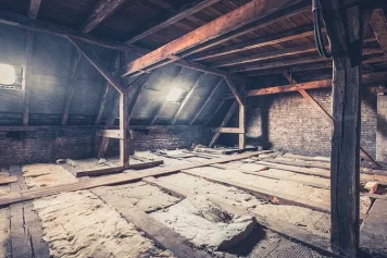 Des combles d’une maison ancienne, avec une charpente en bois apparente, des murs en briques, et un sol en cours de rénovation, laissant entrevoir des traces d’isolation à remplacer.